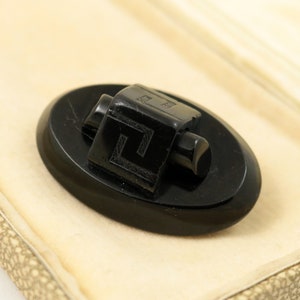 May include: A black oval-shaped button with a black square-shaped design on top. The square design has a Greek key pattern carved into it.