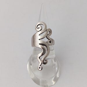 May include: A silver-colored ring with an abstract design featuring swirling, curved lines. The ring is displayed on a clear, cone-shaped stand against a white background. The ring's design has a flowing, organic aesthetic.