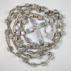 Puede incluir: Un rosario con una cruz central hecha de piedras transparentes de corte cuadrado y facetado. La cadena consiste en un cordón metálico trenzado intercalado con cuentas cuadradas de color gris claro, lisas y con acabado brillante.