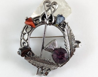 Può includere: Spilla argentata con un design circolare, con un cardo centrale e una grande gemma viola rotonda. La spilla include accenti di piume e gemme in rosso, marrone e blu. Un pezzo decorativo per abbigliamento o accessori.