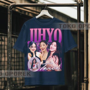 Puede incluir: Camiseta azul marino con un gr&aacute;fico de tres mujeres y el nombre "JIHYO" en morado. Tambi&eacute;n es visible la palabra "Twice". La camiseta est&aacute; colgada en un perchero de metal.