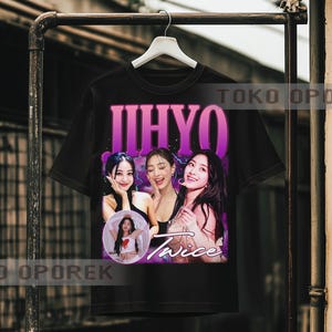 Puede incluir: Camiseta negra con un gr&aacute;fico de tres mujeres, el texto "JIHYO" en morado y "Twice" en blanco. Una imagen m&aacute;s peque&ntilde;a de una mujer est&aacute; en un c&iacute;rculo. La camiseta est&aacute; colgada en un perchero de metal.