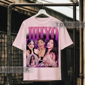 Puede incluir: Una camiseta rosa claro con un gr&aacute;fico de tres mujeres y el nombre "JIHYO" en letras moradas grandes encima de ellas. La palabra "Twice" est&aacute; impresa en cursiva debajo de las mujeres. La camiseta est&aacute; colgada en un perchero de metal.