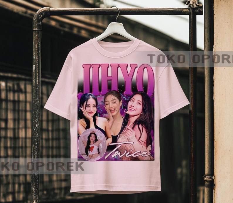 Puede incluir: Una camiseta rosa claro con un gr&aacute;fico de tres mujeres y el nombre "JIHYO" en letras moradas grandes encima de ellas. La palabra "Twice" est&aacute; impresa en cursiva debajo de las mujeres. La camiseta est&aacute; colgada en un perchero de metal.