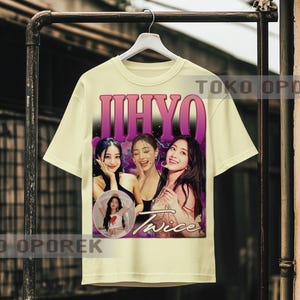 Puede incluir: Camiseta color crema con un gr&aacute;fico de tres mujeres y la palabra "JIHYO" en una fuente degradada p&uacute;rpura. La palabra "Twice" tambi&eacute;n es visible. La camiseta est&aacute; colgada en un estante de metal.