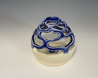 May include: A ceramic candle holder with a cream-colored base and a blue, openwork top. The blue top features a repeating, organic pattern. The candle holder is a decorative item, suitable for home decor.