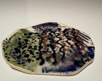 May include: A handmade ceramic plate with an irregular shape. The plate features a colorful abstract design with green, black, red, and blue patterns on a white background. The edges are a light beige color.