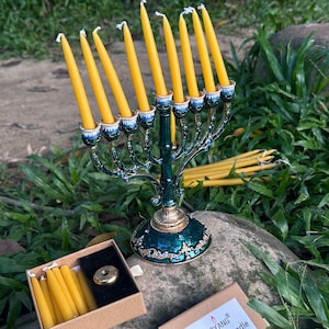 May include: A decorative green and gold menorah with lit yellow candles. Next to it are boxes of yellow beeswax candles. One box is labelled "BOYANG Bee Wax Candle 12 pcs". The menorah sits on a rock with green foliage in the background.