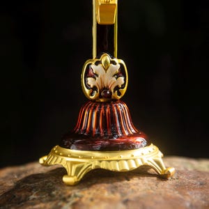 K&ouml;nnte beinhalten: Nahaufnahme eines dekorativen gold- und burgunderfarbenen Kruzifixfu&szlig;es. Der Sockel hat ein detailliertes Design mit einem goldfarbenen floralen Akzent und einem geriffelten, burgunderfarbenen K&ouml;rper. Der Sockel ruht auf einer goldfarbenen, verzierten Plattform mit F&uuml;&szlig;en.