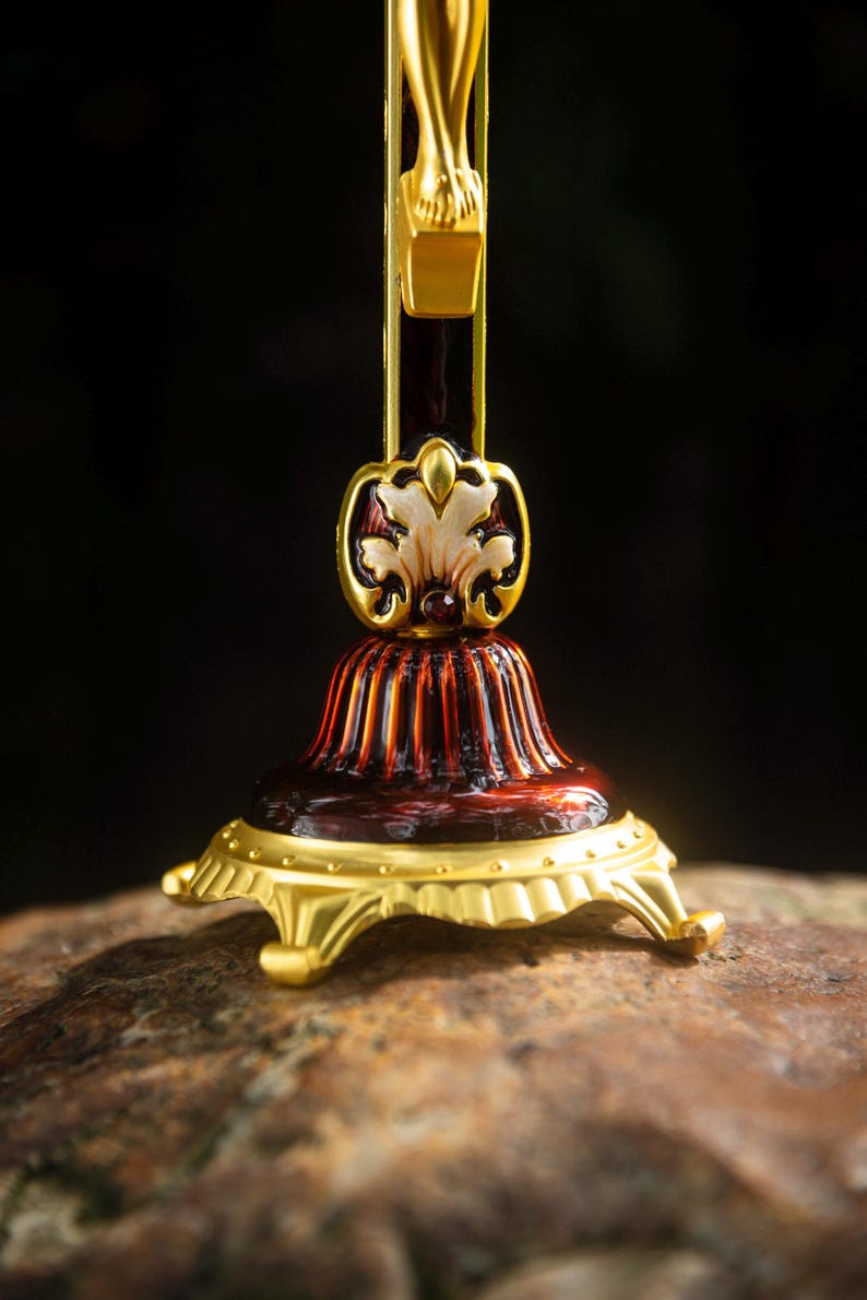 K&ouml;nnte beinhalten: Nahaufnahme eines dekorativen gold- und burgunderfarbenen Kruzifixfu&szlig;es. Der Sockel hat ein detailliertes Design mit einem goldfarbenen floralen Akzent und einem geriffelten, burgunderfarbenen K&ouml;rper. Der Sockel ruht auf einer goldfarbenen, verzierten Plattform mit F&uuml;&szlig;en.