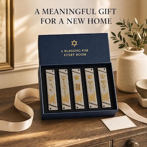 Puede incluir: Una caja de regalo azul oscuro que contiene cinco estuches rectangulares de mezuzá en tono plateado con diseños dorados en relieve que incluyen coronas, menorás y estrellas de David. La tapa incluye el texto A MEANINGFUL GIFT FOR A NEW HOME y A BLESSING FOR EVERY ROOM.