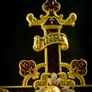 K&ouml;nnte beinhalten: Verziertes goldfarbenes Kruzifix mit einem dunkelroten Kreuz und goldenen Akzenten. Das Kreuz tr&auml;gt die Buchstaben "INRI" auf einem Banner. Die Figur Jesu ist ebenfalls goldfarben, mit detailliertem Design. Der Hintergrund ist schwarz.
