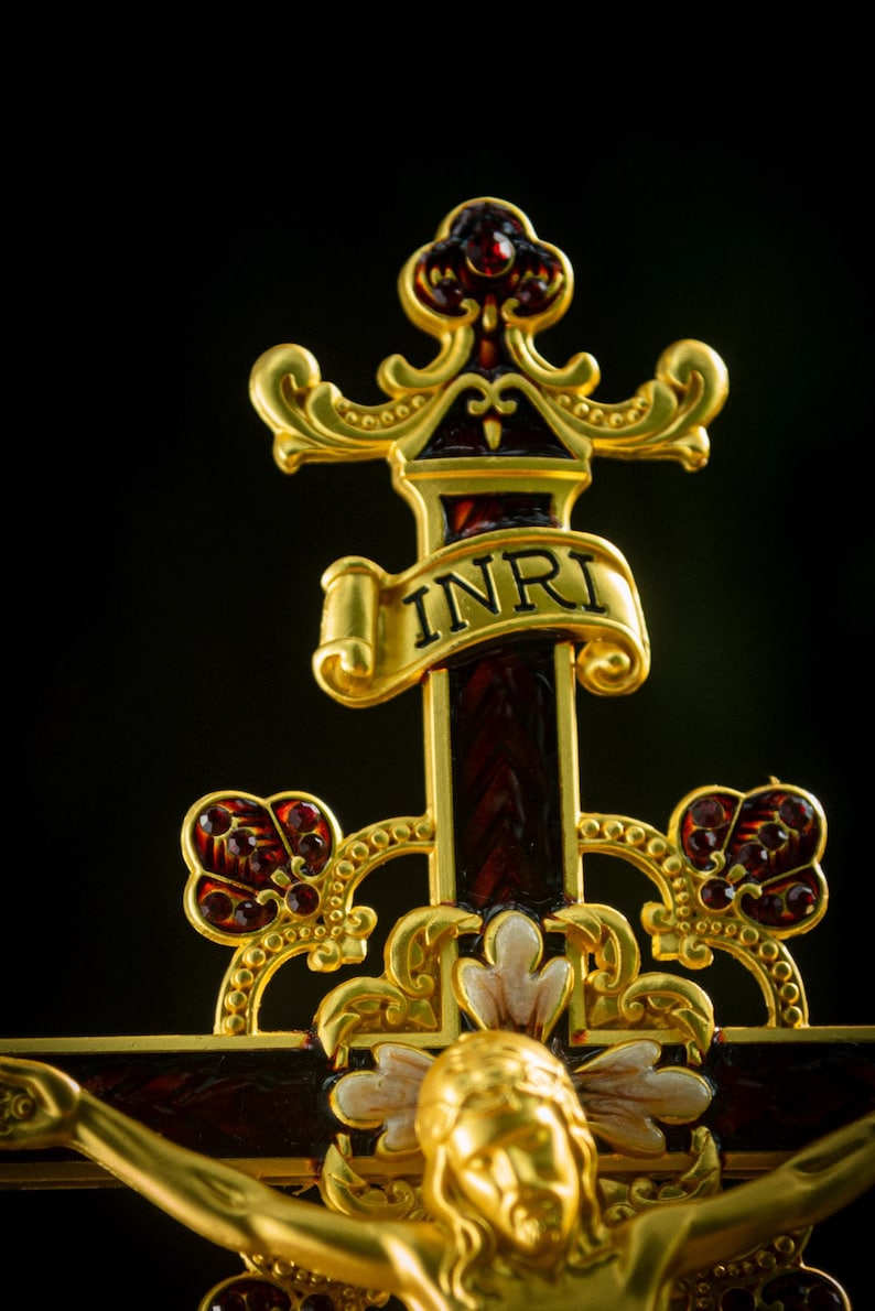K&ouml;nnte beinhalten: Verziertes goldfarbenes Kruzifix mit einem dunkelroten Kreuz und goldenen Akzenten. Das Kreuz tr&auml;gt die Buchstaben "INRI" auf einem Banner. Die Figur Jesu ist ebenfalls goldfarben, mit detailliertem Design. Der Hintergrund ist schwarz.