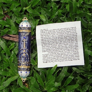 Puede incluir: Un estuche de mezuzá decorativo con un diseño azul y dorado, adornado con piedras preciosas azules. El estuche cilíndrico se coloca sobre hierba verde junto a un papel manuscrito con texto hebreo.