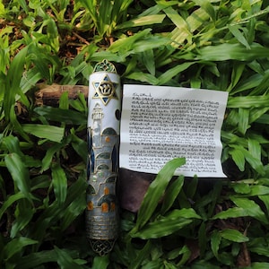 May include: A decorative mezuzah case with a Star of David and architectural design, resting on a bed of green grass. The case is white, blue, and gold. A paper with Hebrew text is next to it.