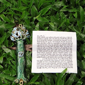 May include: A decorative, tree-shaped pendant with a green and gold stem, adorned with blue leaves and small blue gems. The pendant is set against a backdrop of green grass. A handwritten note is also present.