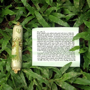 May include: A decorative mezuzah case with a tree design and a scroll with Hebrew text, resting on green grass. The mezuzah case is off-white with gold accents.