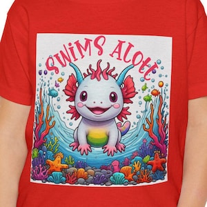 Puede incluir: Camiseta roja con un dibujo animado de ajolote, corales y peces, con el texto "SWIMS ALOT". El ajolote está en el agua. La camiseta la lleva un niño pequeño.