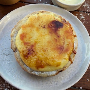 May include: A close-up of a dessert tart with a caramelized custard topping on a white plate. The tart is surrounded by a light-colored crust. Two small bowls of cream are in the background on a wooden surface. The tart is the focal point.