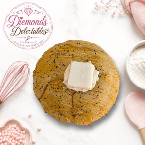 May include: A round, golden-brown biscuit topped with a square pat of white butter, drizzled with honey, and sprinkled with black poppy seeds. The logo for Diemonds Delectables is visible in the upper left corner of the image.