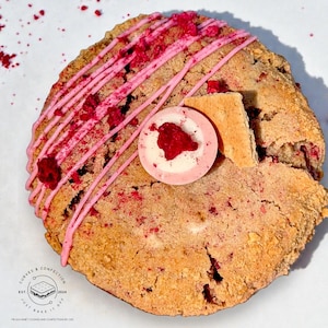 May include: A close-up shot of a large, round cookie with pink icing drizzle and raspberry pieces. The cookie has a light brown color and is topped with a pink and white candy and a piece of honeycomb candy. The cookie is on a white surface.