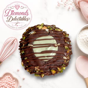 May include: A round chocolate cookie topped with a central pale green cream dollop, drizzled with dark chocolate, sprinkled with chopped green pistachios and gold leaf flakes, featuring the logo Diemonds Delectables Polished to Perfection.