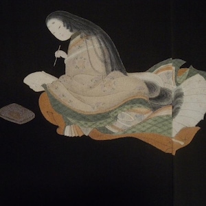 Puede incluir: Una mujer vestida con un kimono japonés tradicional se sienta sobre un fondo negro. Lleva una túnica larga y fluida con intrincados patrones y sostiene un pincel en su mano. El kimono está decorado con motivos florales y tiene una larga cola.
