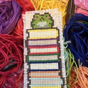 May include: A handmade bookmark featuring a stack of colorful books with a green frog on top. The bookmark is surrounded by various colors of embroidery floss, including red, purple, yellow, and blue.