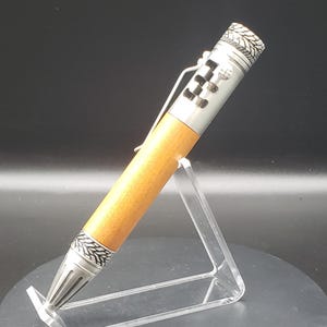 May include: A handcrafted pen with a light brown wooden barrel and silver-colored metal accents. The pen features a clip and decorative patterns on the metal components. The pen is displayed on a clear acrylic stand.