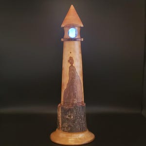 May include: A wooden lighthouse lamp with a light brown finish. The lamp features a conical roof, a circular window with a blue light, and a base made of a tree trunk. The lighthouse is approximately 12 inches tall.