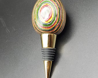 May include: A decorative bottle stopper with a colorful, layered wooden egg-shaped top. The top features concentric rings of various colors. It has a gold-colored metal base and a black rubber seal. The stopper is designed to fit into a bottle.