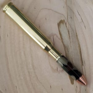 Könnte beinhalten: Ein goldfarbener Stift mit kupferfarbener Spitze und einem Griff mit Camouflage-Muster. Der Stiftkörper hat einen metallischen Glanz, und das Design deutet auf ein einzigartiges, handgefertigtes Schreibgerät hin. Der Stift liegt auf einer hellen Holzoberfläche.