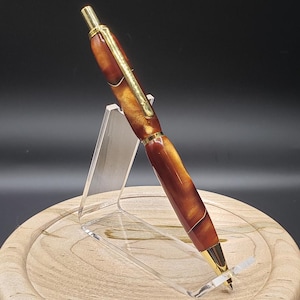 May include: A brown and gold pen with a swirl pattern on the barrel. The pen is on a clear acrylic stand.