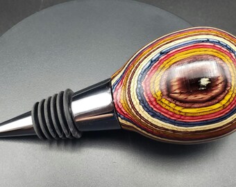 May include: A decorative bottle stopper with a colorful, layered wood design. The stopper features a polished, oval-shaped top with concentric rings of red, yellow, blue, and brown. It has a stainless steel collar and a black rubber seal.