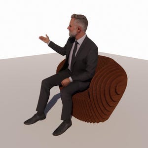 May include: A man in a dark suit sits in a brown, layered wooden chair. The chair has a unique, spiral design. The man is gesturing with his right hand, as if presenting or speaking.