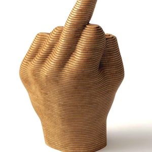 Può includere: Scultura in legno di una mano con il dito medio rivolto verso l'alto. La mano è costruita con legno a strati, con linee orizzontali visibili. La scultura è di colore marrone chiaro e si erge su uno sfondo bianco.