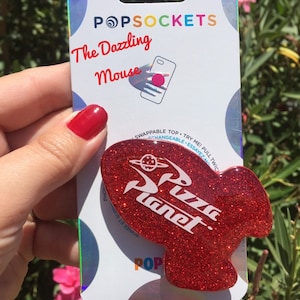 May include: Red glitter phone grip with a white "Pizza Planet" logo. The grip is shaped like a rocket ship and is packaged with a PopSockets brand card.