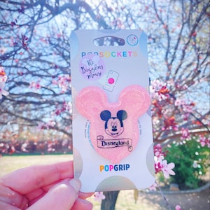 May include: Pink glitter phone grip with a Mickey Mouse head design. The grip features a black and white image of Mickey Mouse with the word "Disneyland" on a scroll. The packaging says "The Dazzling Mouse" and "PopGrip".