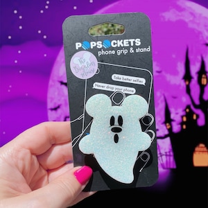 May include: A white glitter ghost PopSocket phone grip and stand with a black background. The packaging features the text "PopSockets", "Phone grip & stand", "The Dazzling Mouse", "Take better selfies", and "Never drop your phone".