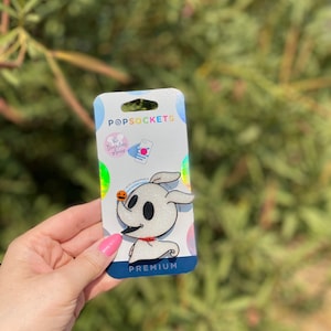 May include: A white and silver PopSockets phone grip featuring a cartoon character from The Nightmare Before Christmas. The character is Zero, the ghost dog, holding a small orange pumpkin. The text "PREMIUM" is printed on the packaging.