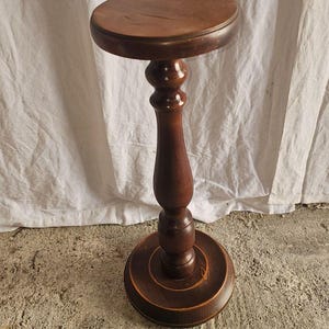 Può includere: Un supporto a piedistallo in legno marrone scuro con piano circolare, colonna centrale tornita con dettagli decorativi bombati e una base circolare ampia e stabile. La venatura del legno è visibile su tutta la struttura dell'oggetto.