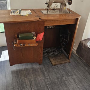 Può includere: Una macchina da cucire Singer vintage montata in un mobile in legno con tavolo pieghevole. La macchina è beige e dotata di lampada. Il mobile presenta un ripiano interno con accessori per cucire e un pedale poggiato sul pavimento.