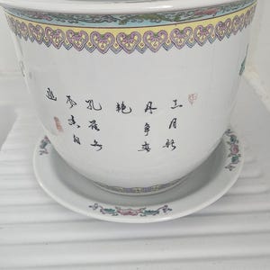 Peut inclure: Pot de fleurs en céramique blanche avec soucoupe assortie. Le pot présente un motif floral et géométrique décoratif sur le bord et sur la soucoupe. Des caractères chinois sont écrits sur le côté du pot.
