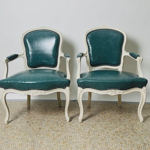 May include: Two antique armchairs with teal leather seats and backs. The chairs have a cream-colored wooden frame with curved legs and armrests. The seats and backs are trimmed with black studs.