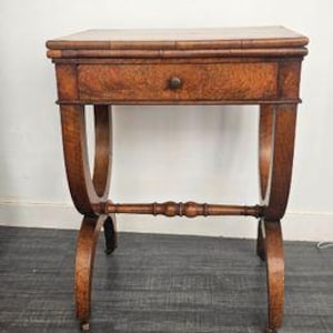 Può includere: Un tavolino in legno marrone con piano rettangolare, un cassetto singolo con un piccolo pomello rotondo e gambe curve collegate da un traversino orizzontale centrale. Il tavolo presenta una venatura del legno visibile e poggia su piccole rotelle.