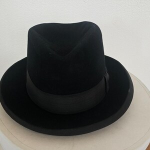 Könnte beinhalten: Ein schwarzer Fedora-Hut mit eingedrückter Krone und breiter Krempe, der mit einem passenden schwarzen Ripsband um die Basis versehen ist. Der Hut liegt auf einer weißen zylindrischen Schachtel mit dem Teiltext CHAPEAUX OTSCH PARIS.