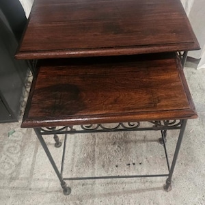 Op de afbeelding: Een set van twee bijzettafels met rechthoekige donkerbruine houten bladen met zichtbare houtnerven. De tafels worden ondersteund door een zwart metalen frame met decoratieve kruldetails langs de bovenzijden en vier dunne poten.