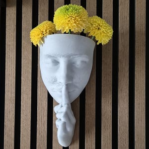 Può includere: Una scultura in gesso bianco di un viso con un dito sulle labbra, come per dire 'shh'. La scultura è usata come vaso e ha fiori gialli che crescono dalla parte superiore.