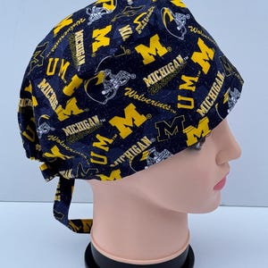 May include: A navy blue and yellow patterned scrub cap featuring the text "Michigan Wolverines" and the letter "M". The cap has a tie in the back and is designed to be worn on the head.