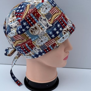 May include: A patterned scrub cap featuring a patriotic design with American flags, stars, and text. The cap is predominantly blue, red, and white, with a tie closure at the back. The text includes phrases like "Liberty and Justice for All" and "One Nation Under God."